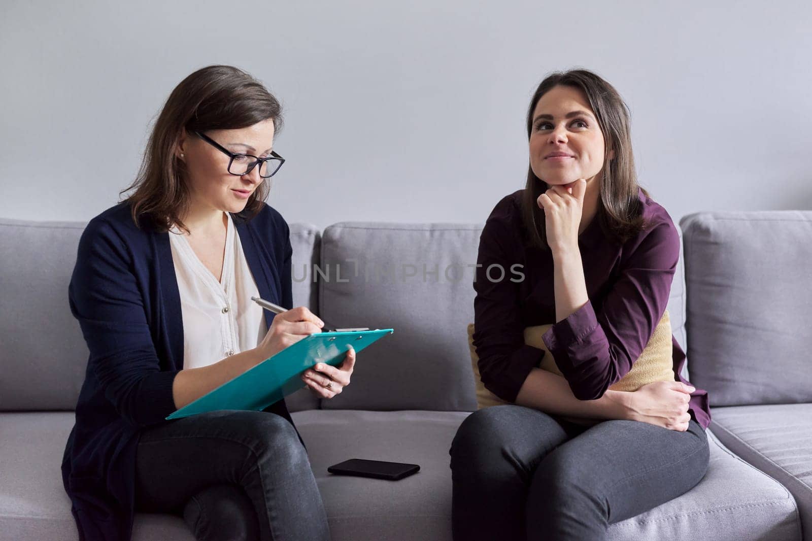 Social worker, psychologist talking to young woman in office. Women sitting on couch, counselor making notes, listening to patient. Psychology, mental health, social difficulties of young people