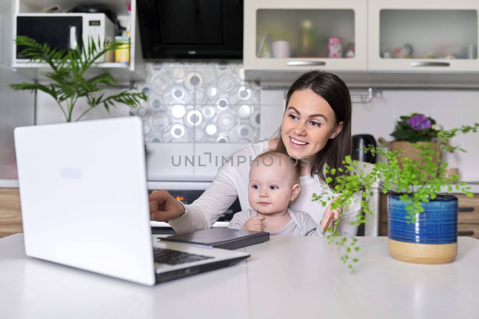 Portrait of mom and baby son sitting at home in kitchen looking in laptop screen. Working at home, blogging, vlog, business, rest and leisure, e-learning for young mother, online shopping