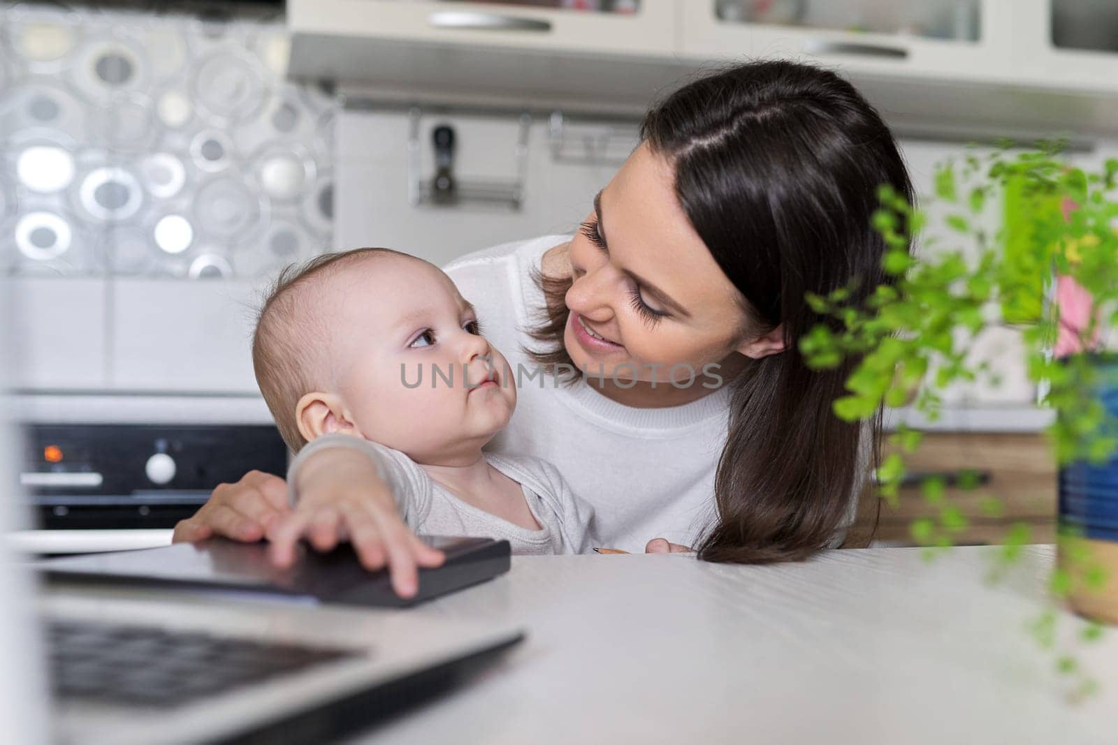 Portrait of mom and baby son sitting at home in kitchen looking in laptop screen. Working at home, blogging, vlog, business, rest and leisure, e-learning for young mother, online shopping