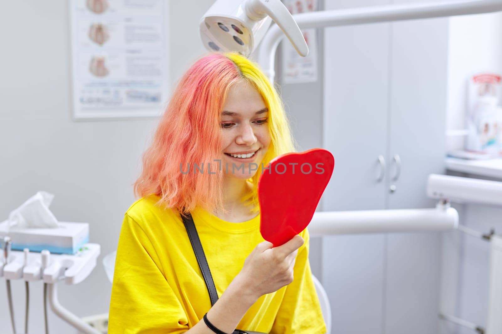 Happy patient beautiful teenage girl looking at her healthy white teeth in mirror smiling while sitting in dental chair in clinic