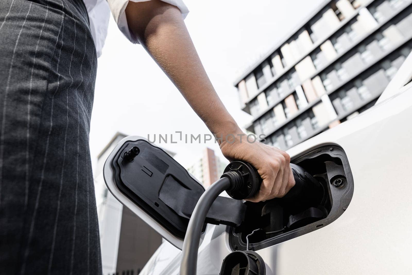 Focus EV charger plug and electric car at public charging station with blur progressive businesswoman holding charger and apartment condo building in background. Eco friendly electric vehicle concept.