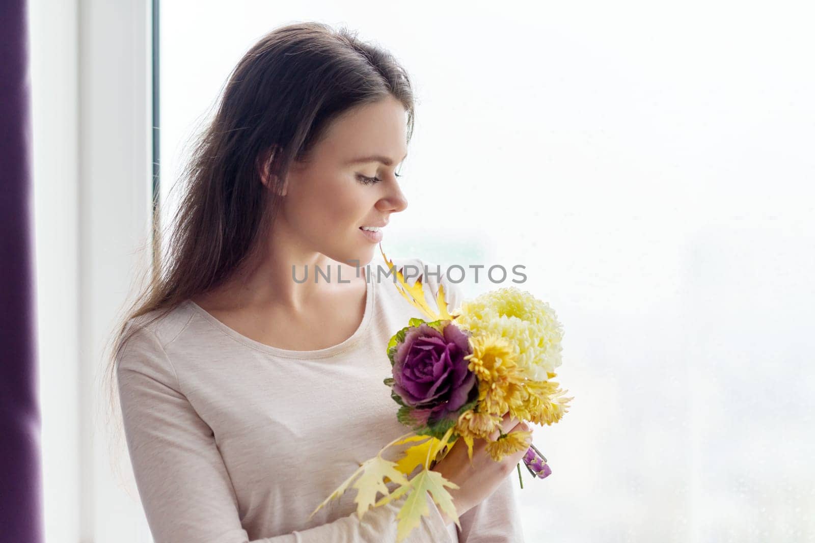 Autumn background, young beautiful woman with bouquet of yellow fallen leaves and autumn flowers looking in cloudy rainy window, copy space for text