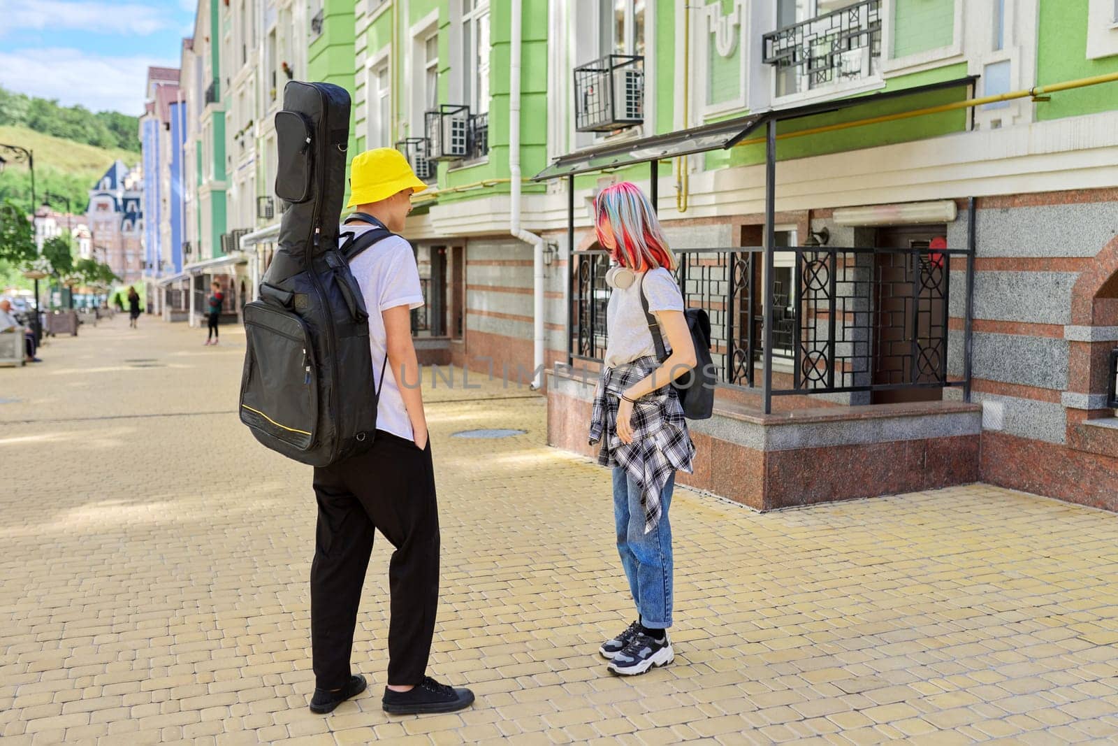 Trendy teenage hipsters boy and girl walking talking on city street, boy with guitar in case. Youth, lifestyle, creativity, communication, friendship concept
