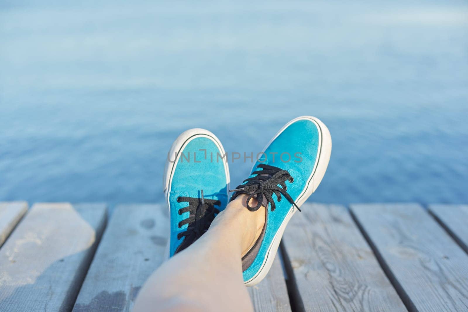 Womans legs in blue sneakers on wooden pier, river lake pond background for copy space. Vacation, nature, lifestyle, people concept