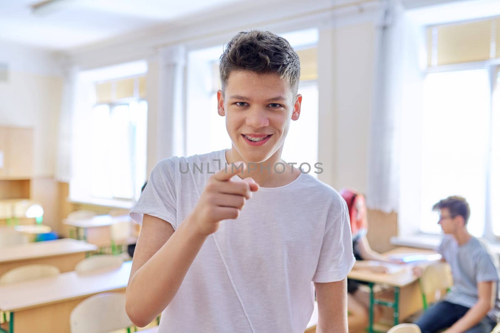 Smart teenager student male taking exam, answers lesson, talking looking at camera, classroom with study students background. High school, education, youth concept