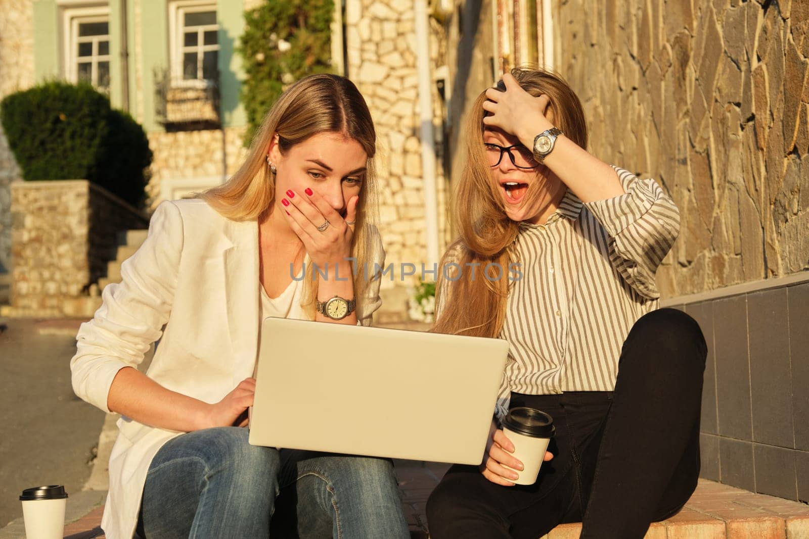 Two young beautiful women having fun looking into the laptop monitor, business females sitting on steps in city street, university students laughing resting