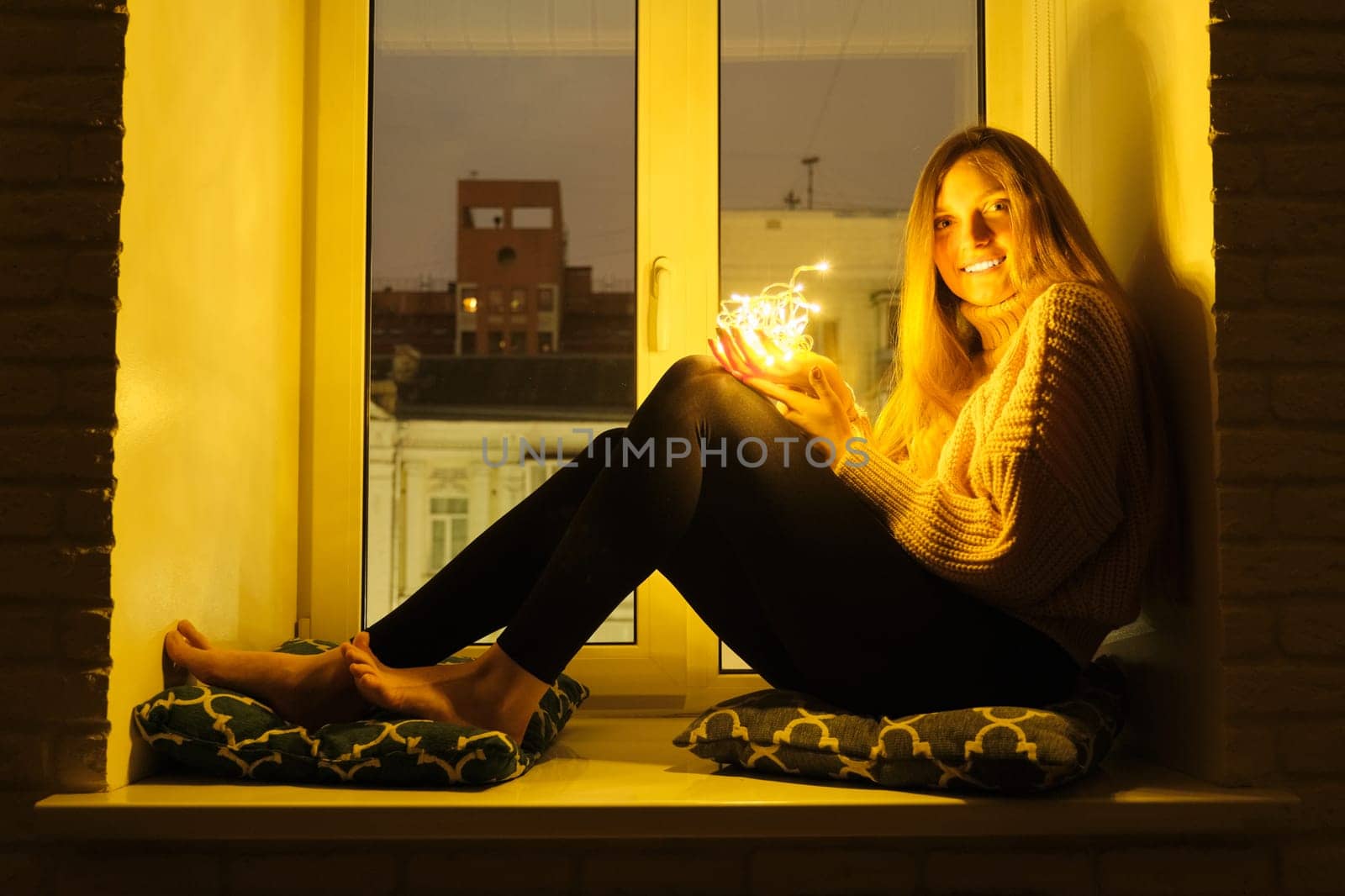 Winter portrait of young beautiful woman sitting near window on windowsill, night festive city background, sparkling garland lights in hands of girl