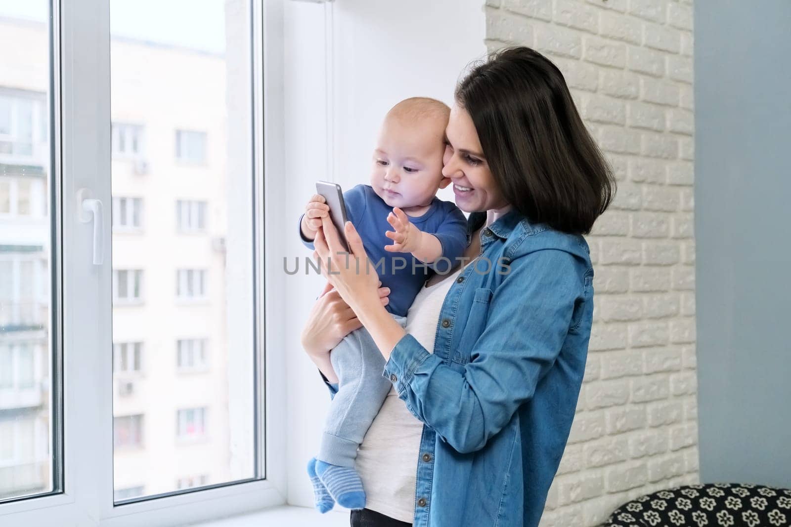 Young mother and son toddler baby in their arms are looking at smartphone screen, video call, online communication. Happy family at home, near window, winter autumn season