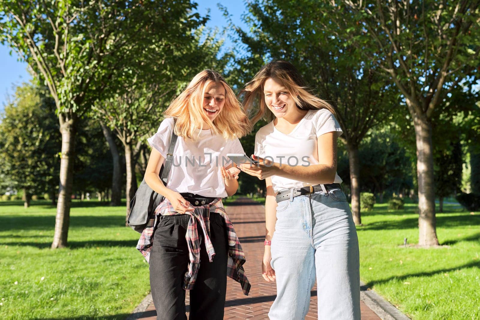 Two happy talking walking teenage girls looking at smartphone screen, college students walking in park laughing having fun. Youth teens, lifestyle, students concept