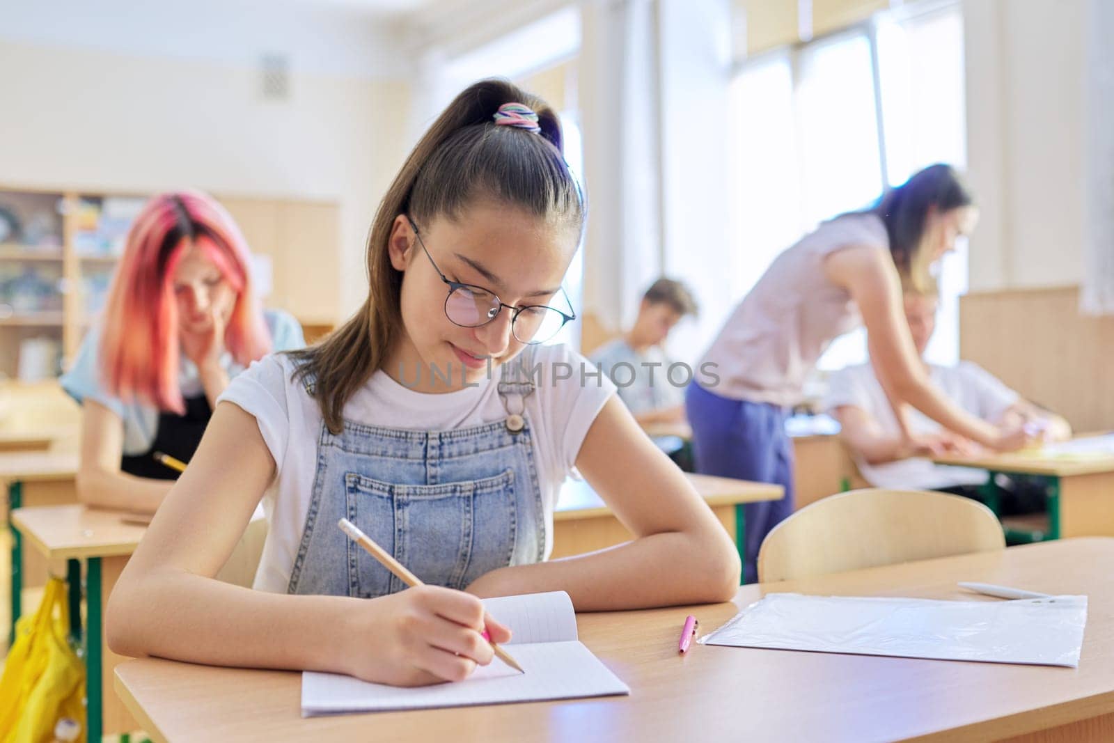 Lesson in class of teenage children, in front girl 13, 14 years old sitting at desk writing in notebook. Education, school, college, schoolchildren concept