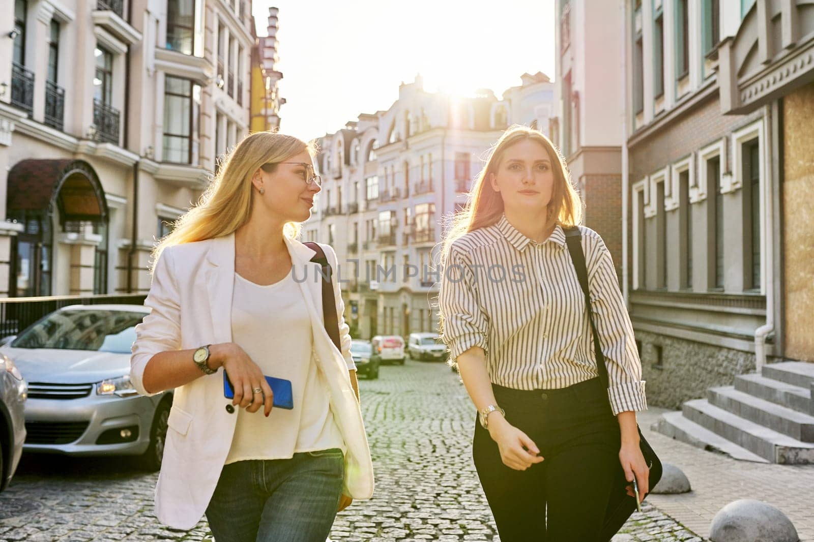 Two young beautiful happy women university students walking along city street by VH-studio