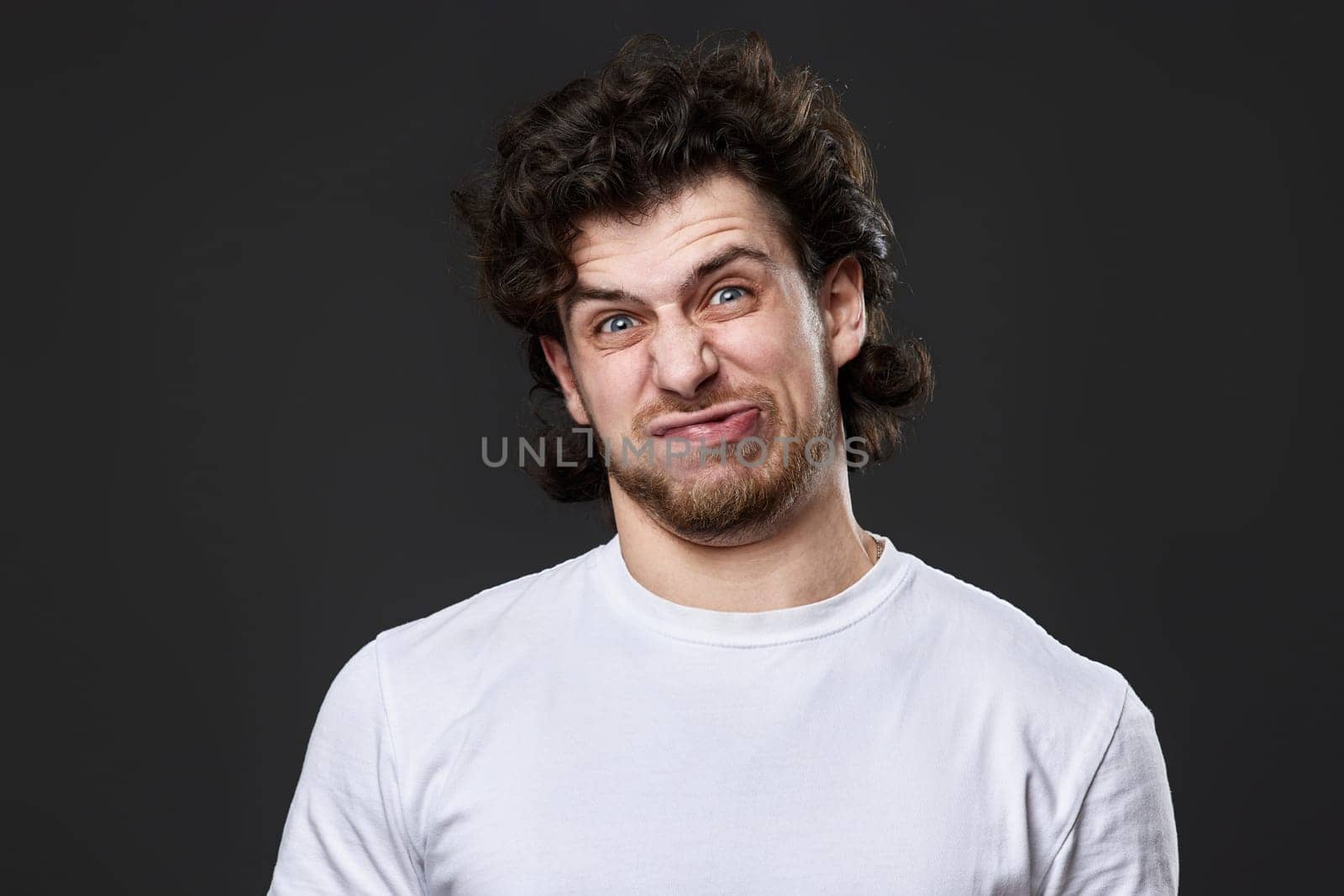 portrait of young funny curly man in white t-shirt grimacing on dark gray background