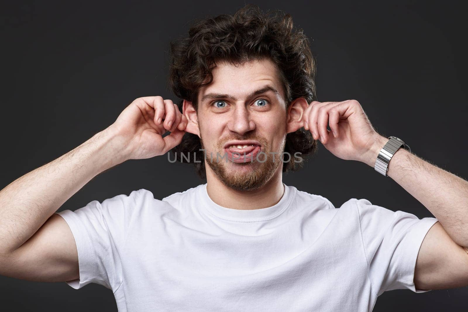 portrait of young funny curly man in white t-shirt grimacing on dark gray background