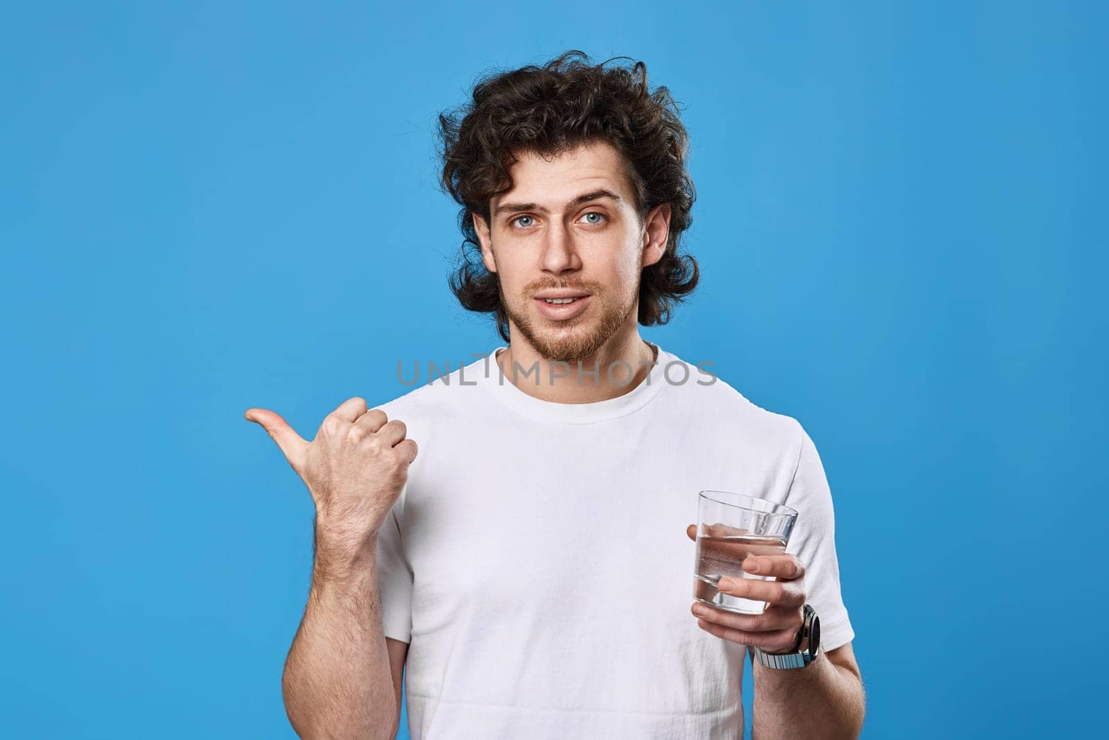 handsome bearded man in white t-shirt with drinking glass of water pointing to the side on blue background
