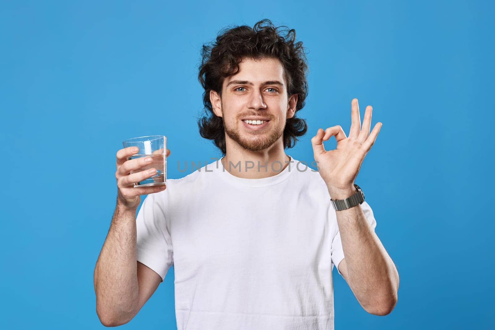 Young handsome man in white t-shirt with drinking glass of water showing Ok gesture on blue background