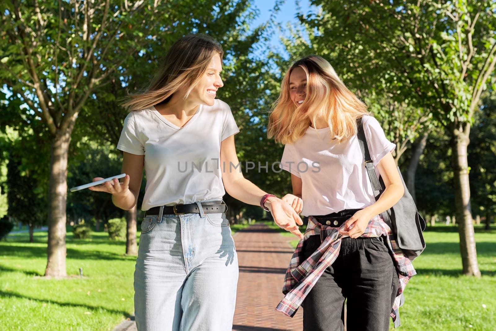 Two beautiful happy girls teenagers 17, 18 years old walking together in park, girls laughing talking having fun on sunny summer day