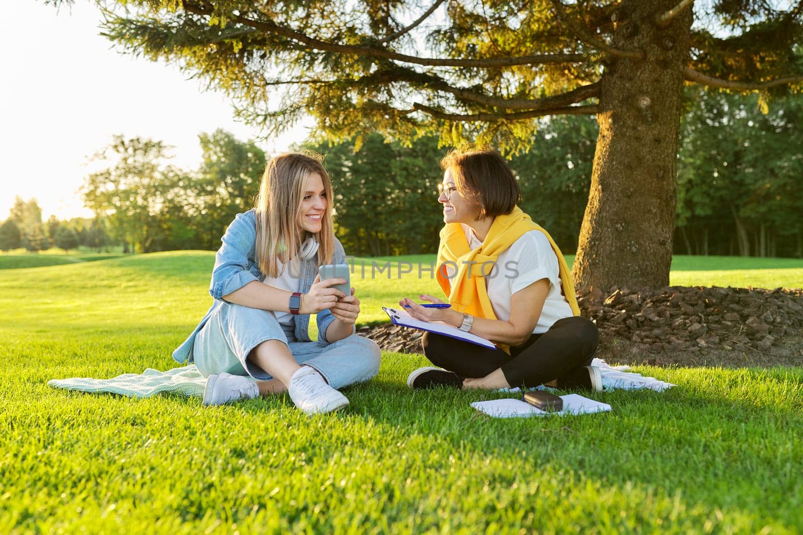 Outdoor meeting of teenage girl and woman of psychologist social worker, women talking sitting on green lawn in park, comfortable environment for talking with young people, mental help consultation