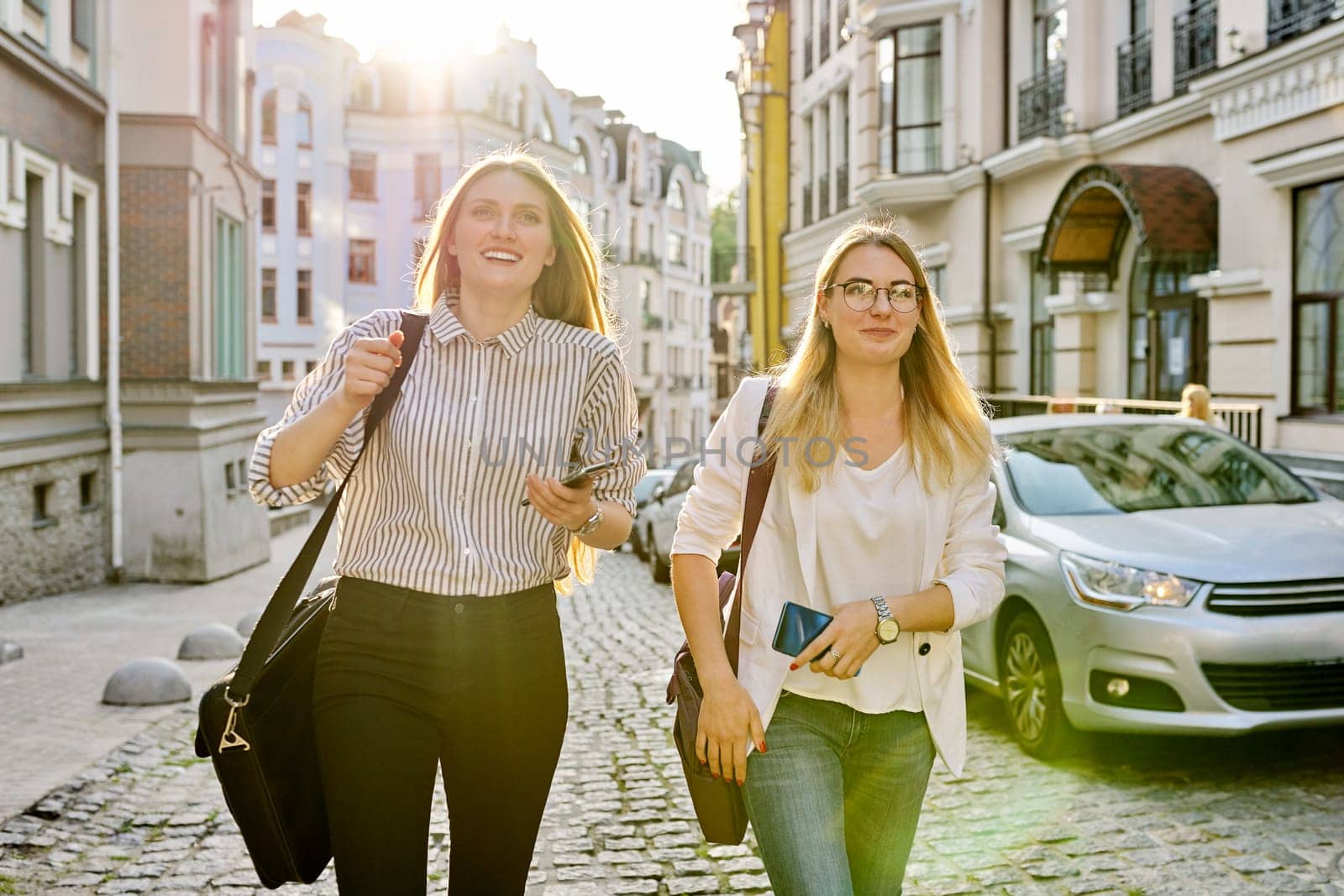 Two young beautiful happy women university students walking along city street, women smiling laughing looking forward