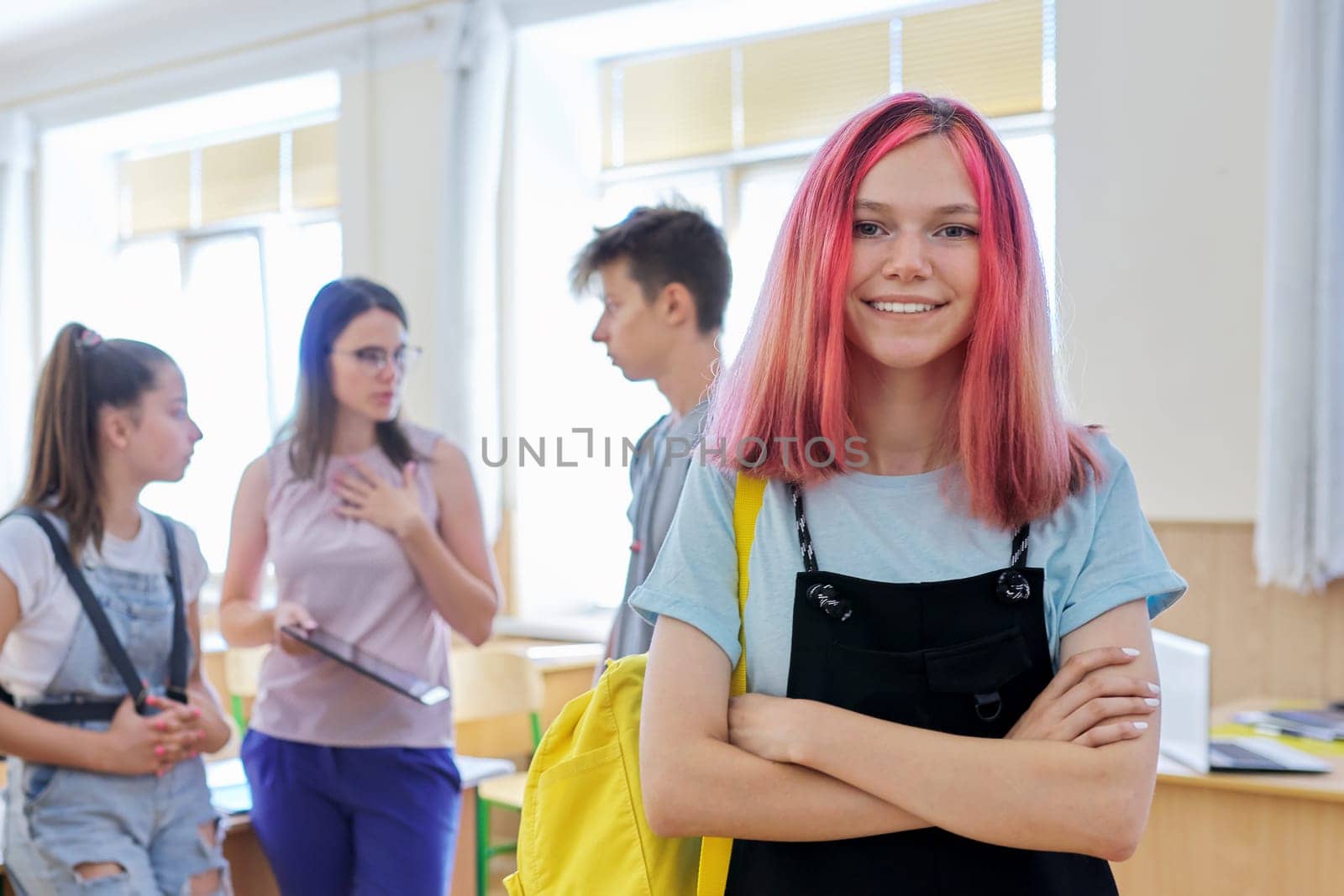 Portrait of smiling teenage girl in class on break. Background group of talking teenage classmates and teacher. School, college, study, youth concept