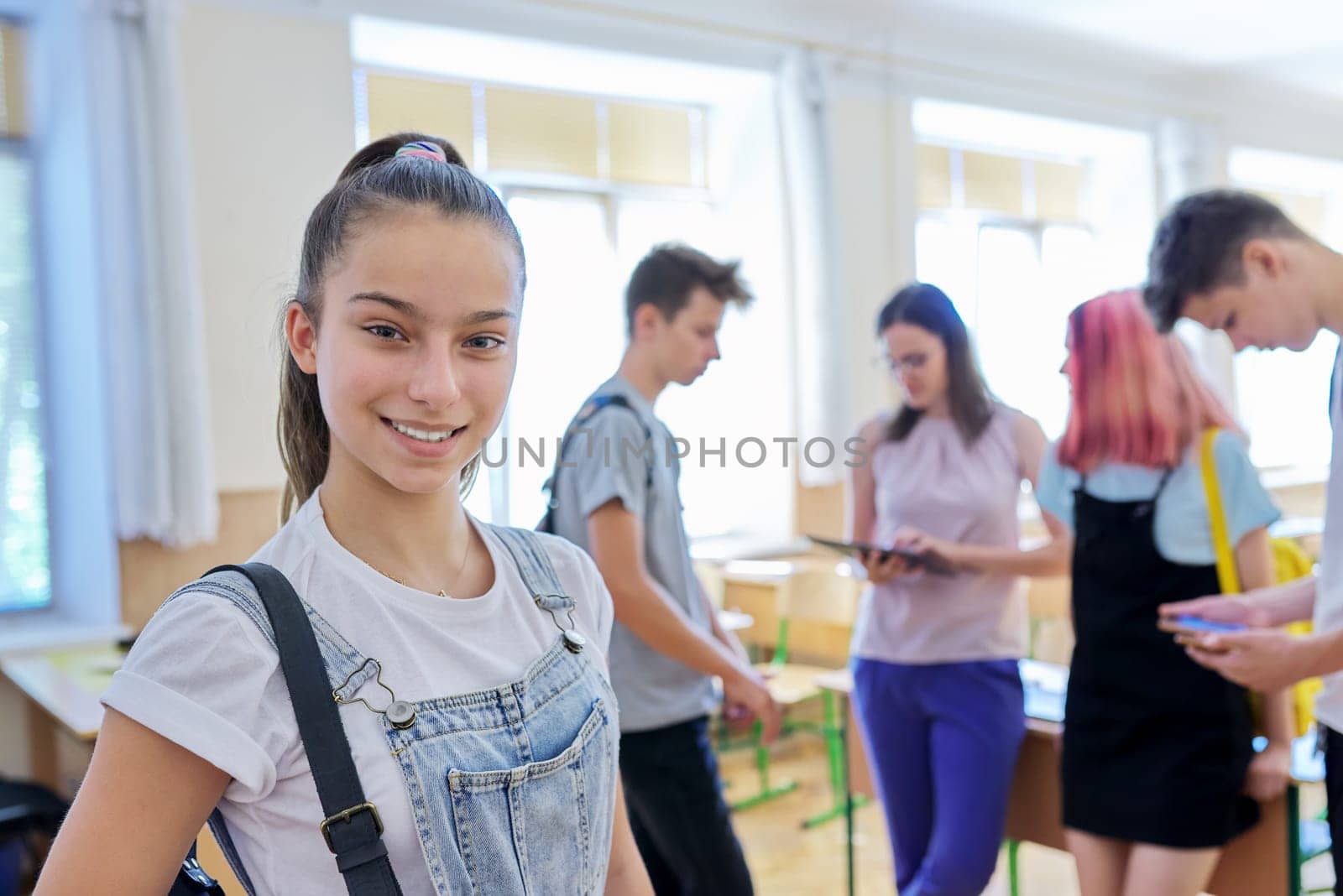 Portrait of smiling teenage girl in class on break. Background group of talking teenage classmates and teacher. School, college, study, youth concept
