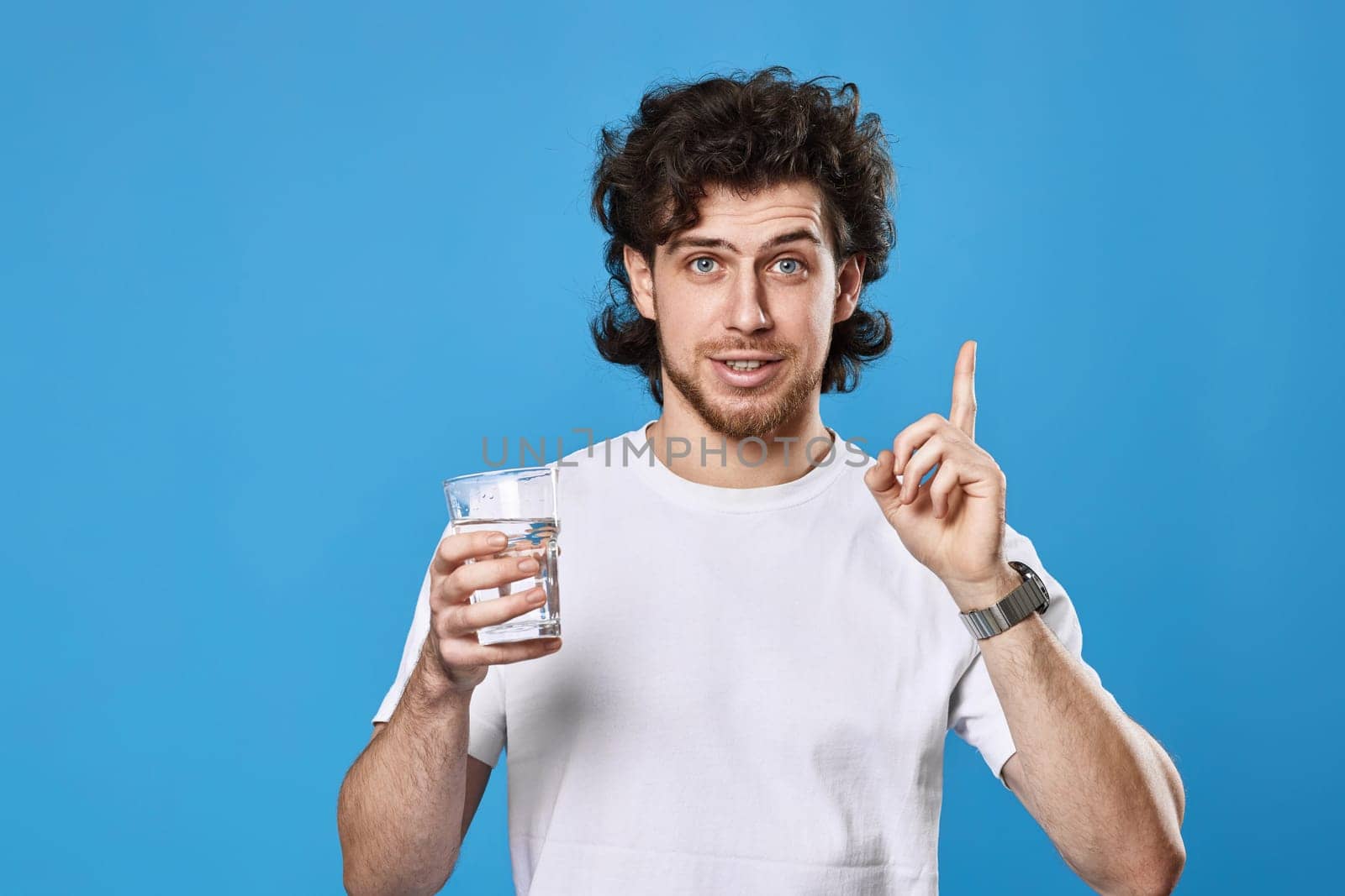 Young curly man in white t-shirt with drinking glass of water pointing up on blue background