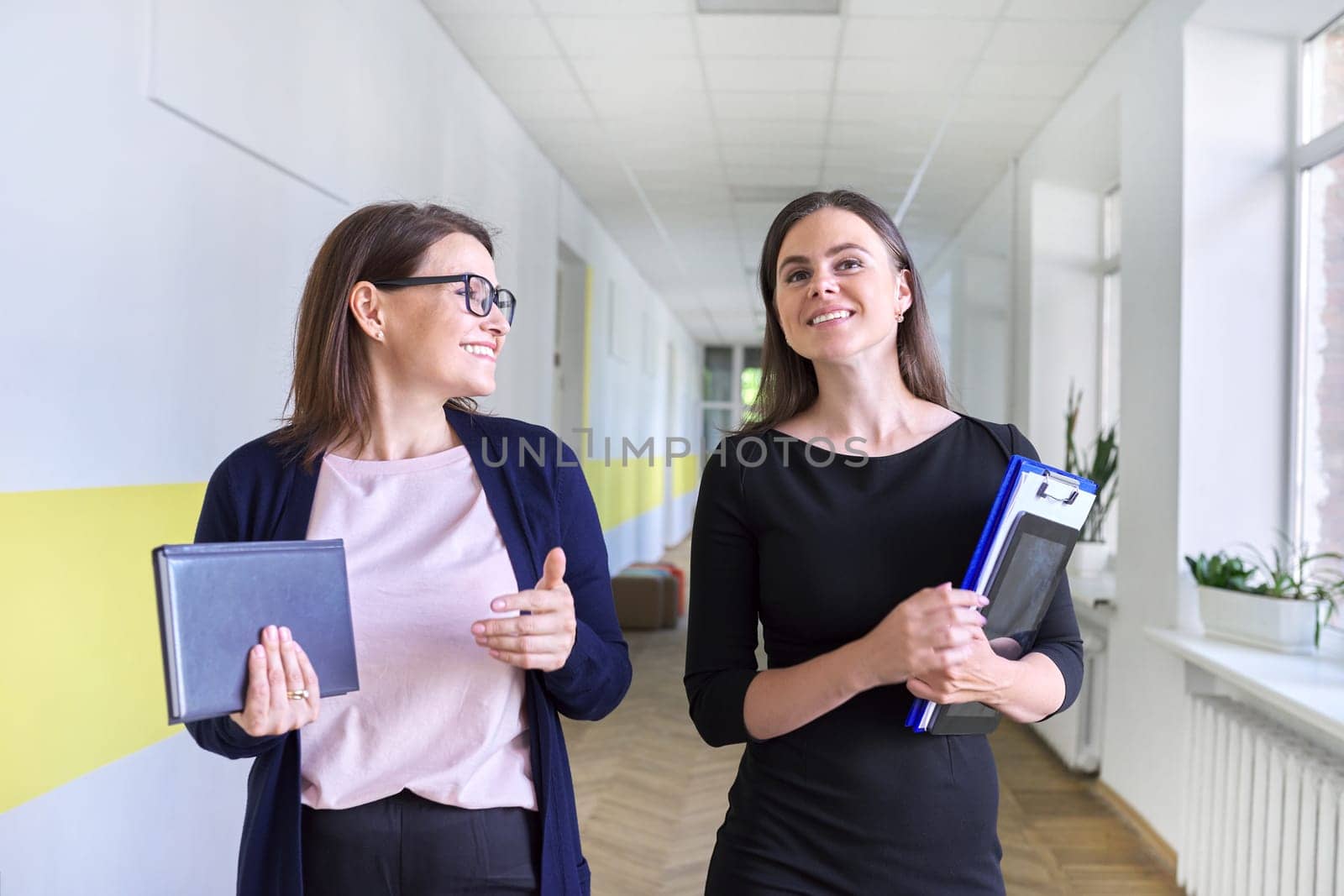 Two colleagues of businesswoman, teacher walking and talking on corridor of office, school. Positive smiling young and middle aged females, business education professions office workers concept
