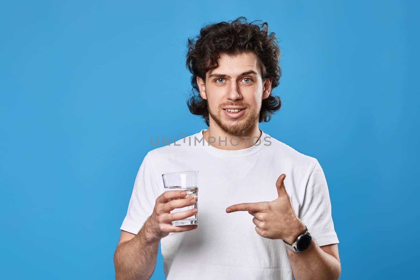 young positive man in white t-shirt showing drinking glass of water on blue background
