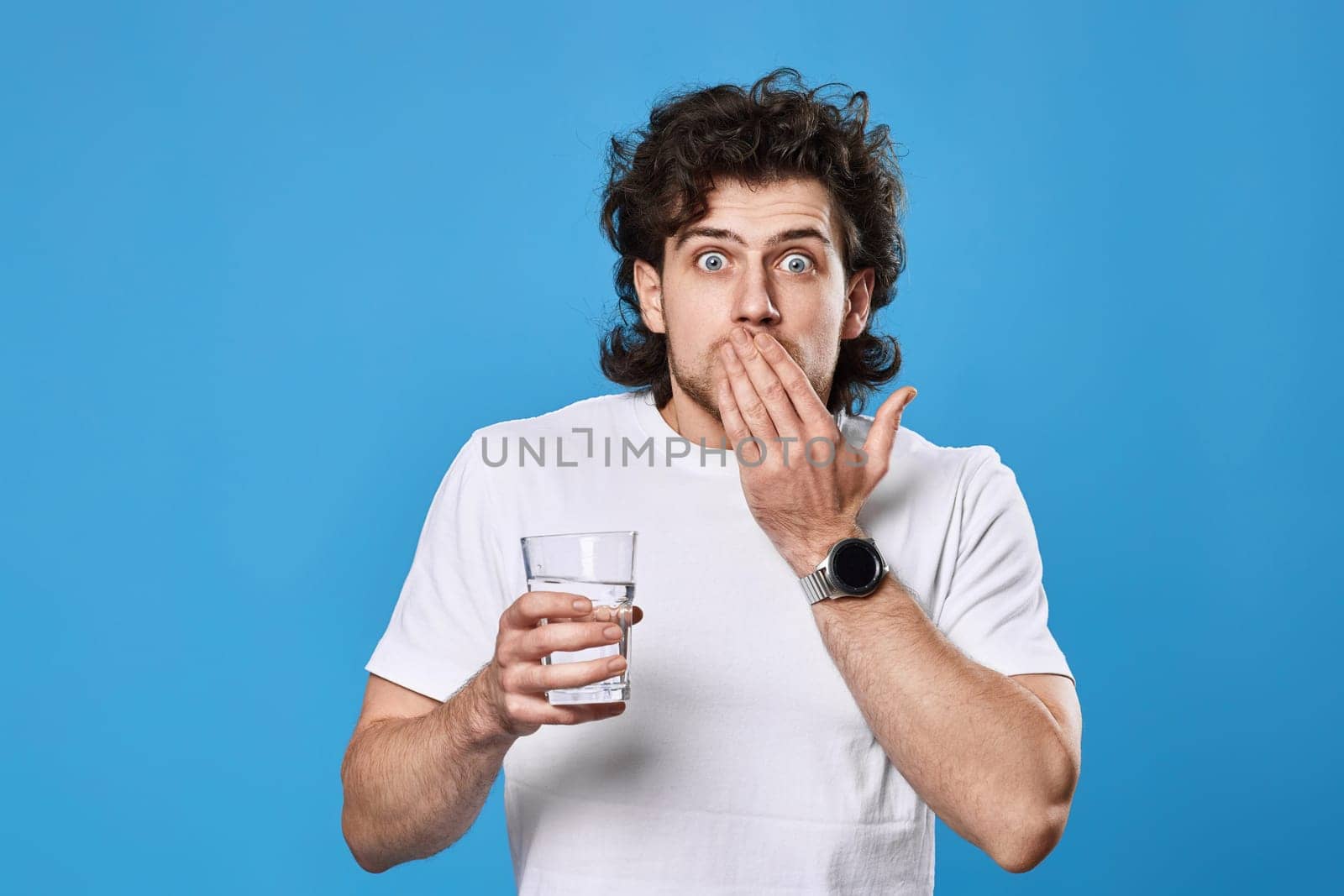 Young surprised handsome man with drinking glass of water on blue background