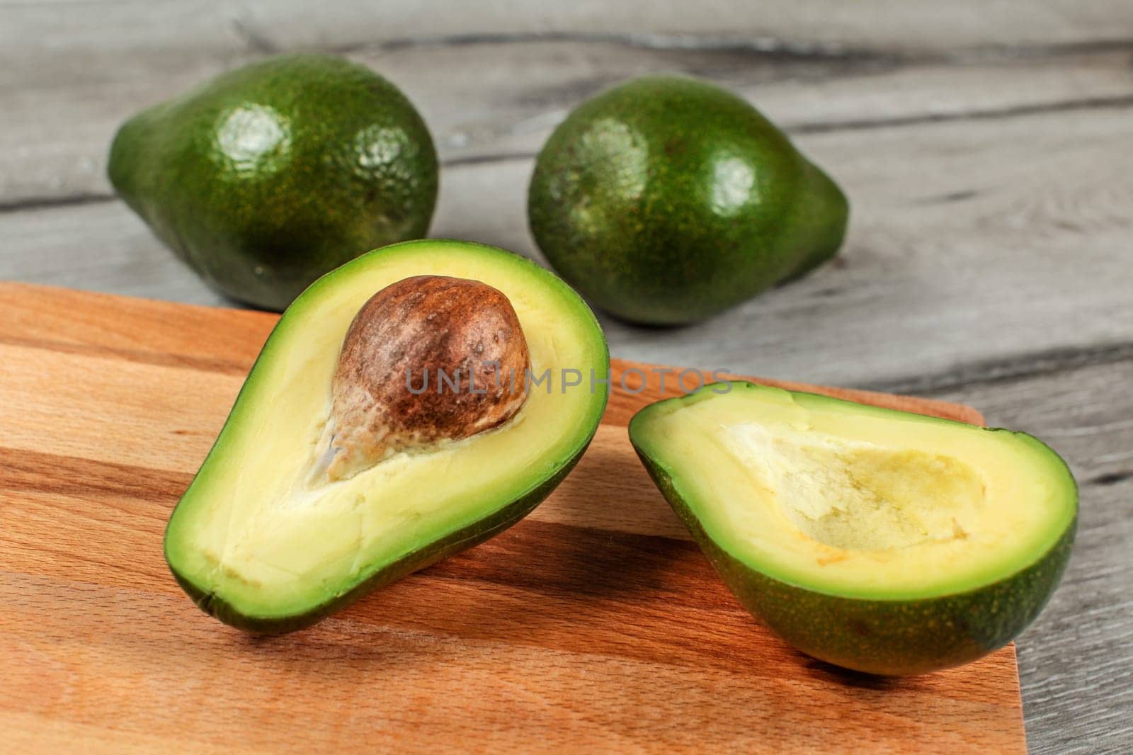 Avocado cut in half, seed visible, on wooden chopping board, two more whole pears in background.