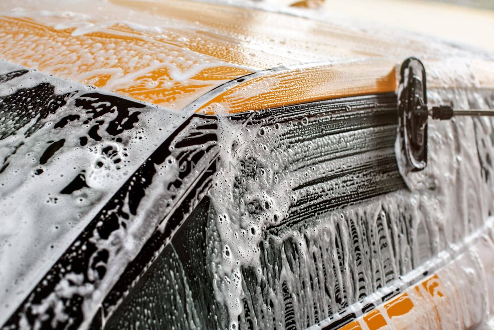 Yellow car washed in self service carwash, brush leaving strokes in white shampoo and foam on side window glass.