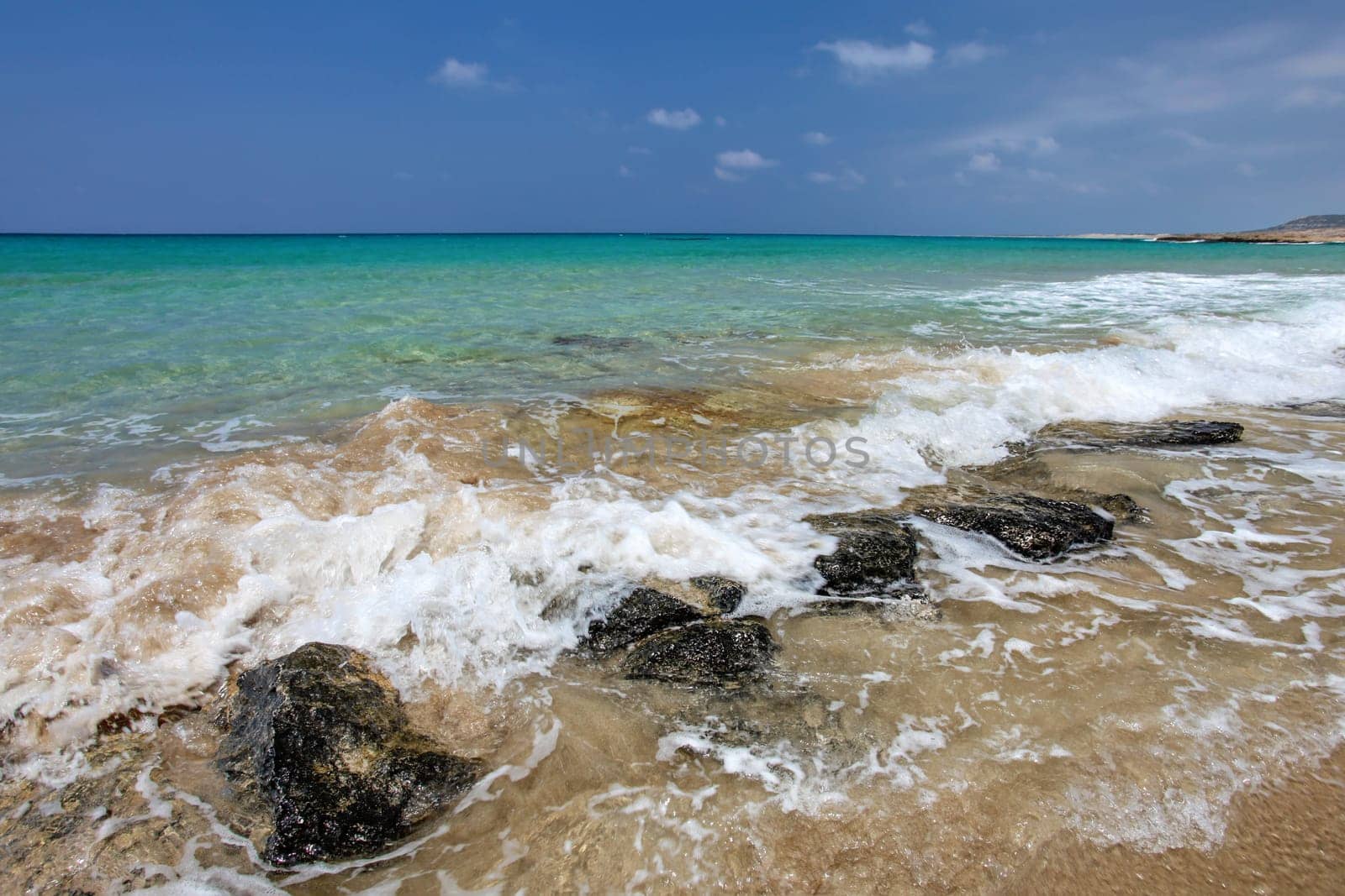 Small sea waves crushing on black rocks at sandy beach, beautiful blue crystal clear water in background. Karpass, Northern Cyprus