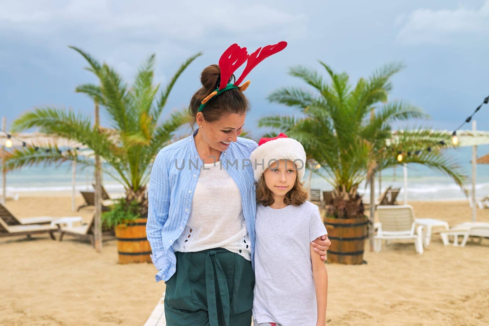 Happy mom and daughter child in Santa hat having fun walking along the beach embracing. Christmas and New Year holidays together with family at tropical resort, sea ocean palm trees background