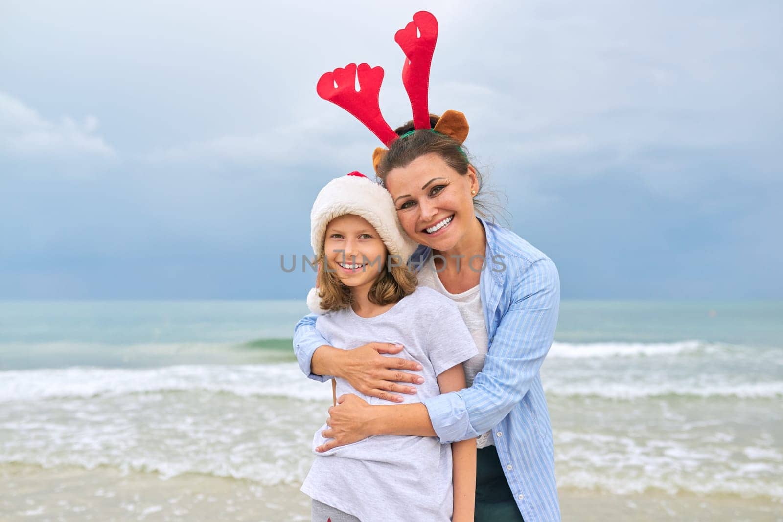 Portrait of happy embracing mom and daughter, parent and child celebrating Christmas and New Years together. Sea ocean beach sky background, family holidays travel concept