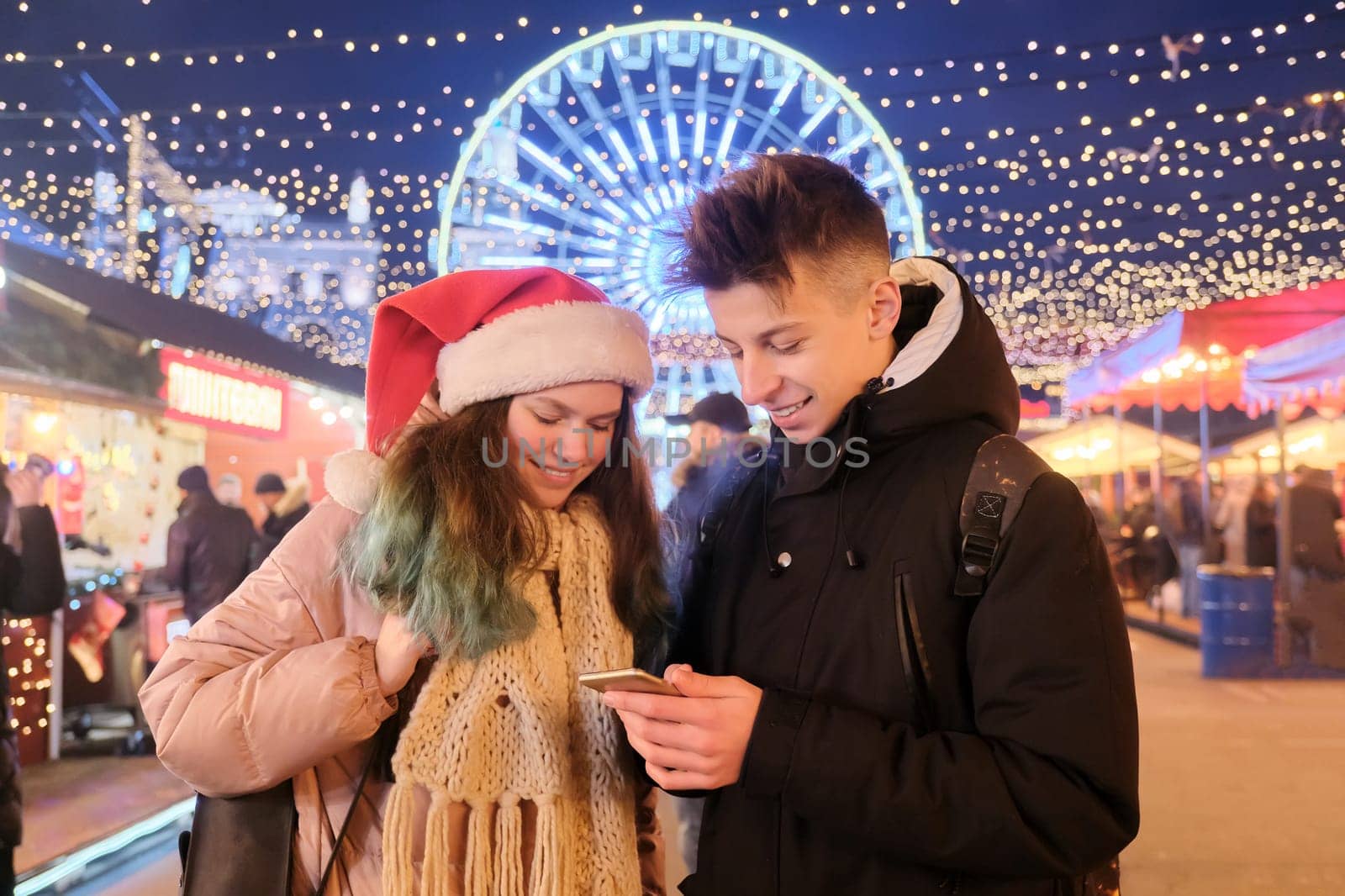Teenagers boy and girl at the Christmas market, in Santa hat, with a smartphone. Lights, bright garlands of evening city, ferris wheel background