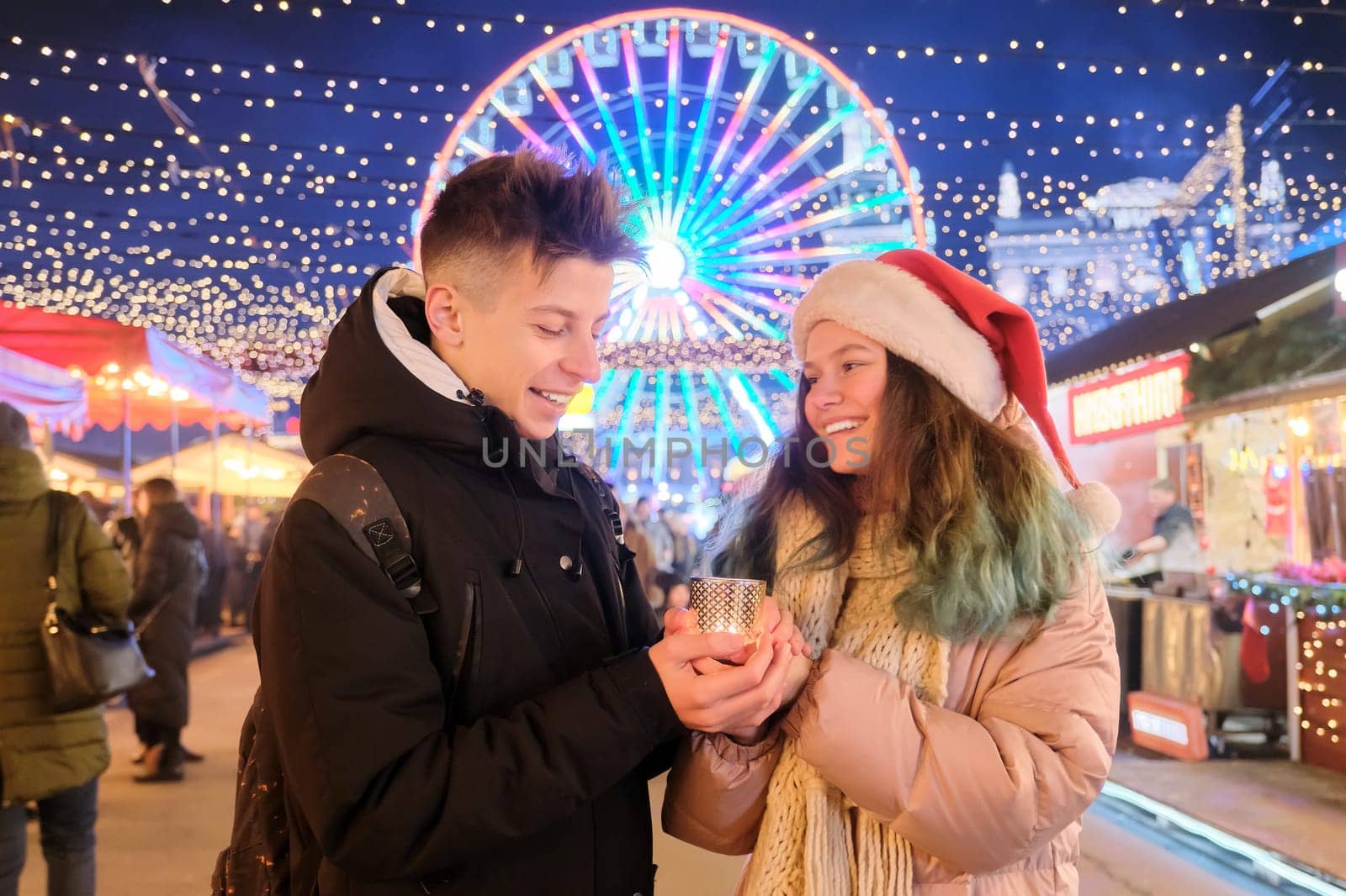 Happy teenagers couple boy and girl in Santa hat at Christmas market warming hands with candle. Lights, bright garlands of evening city, ferris wheel background