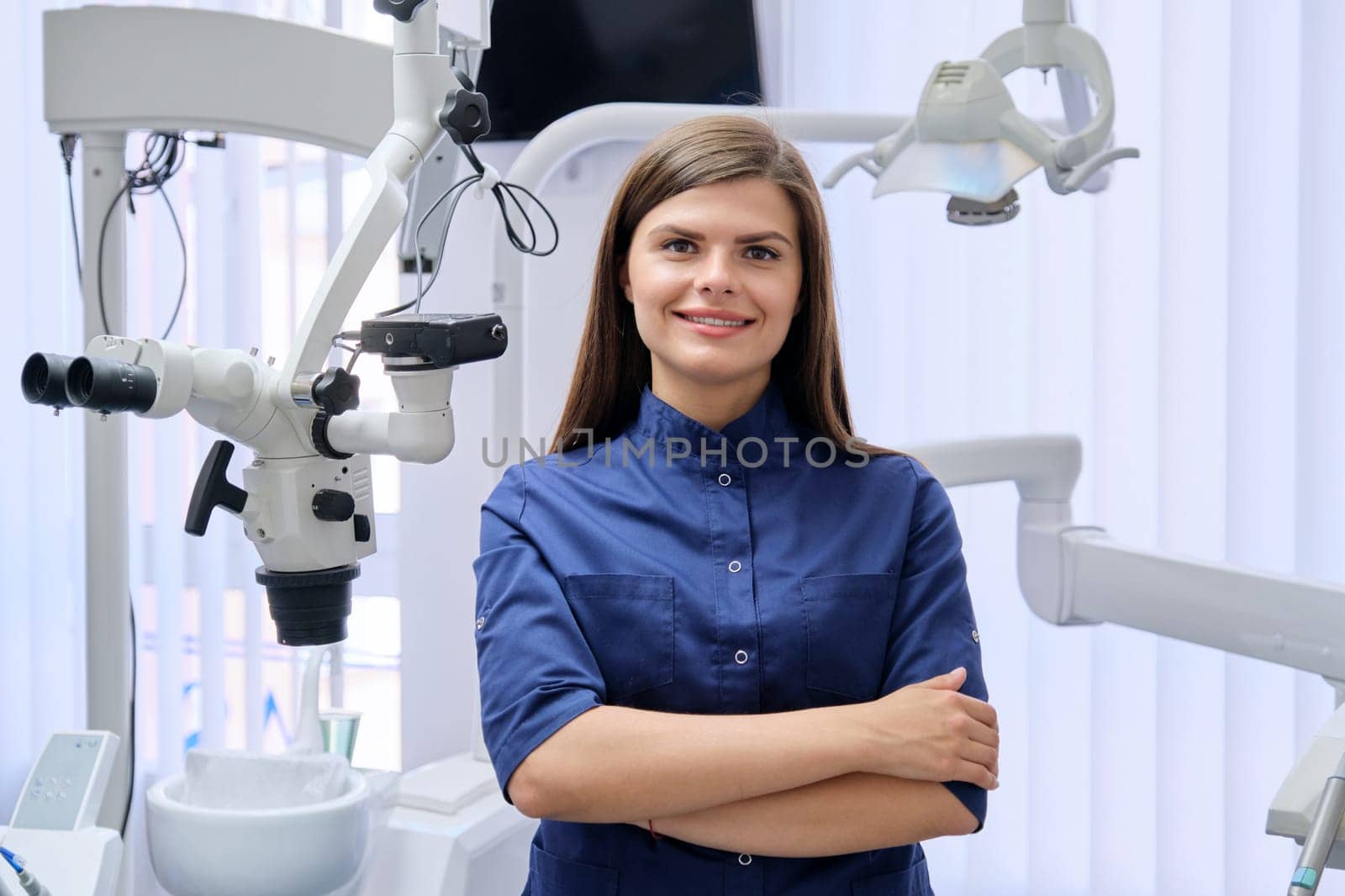 Portrait of young confident female dentist doctor with folded hands in dental office. Medicine, dentistry and health care concept