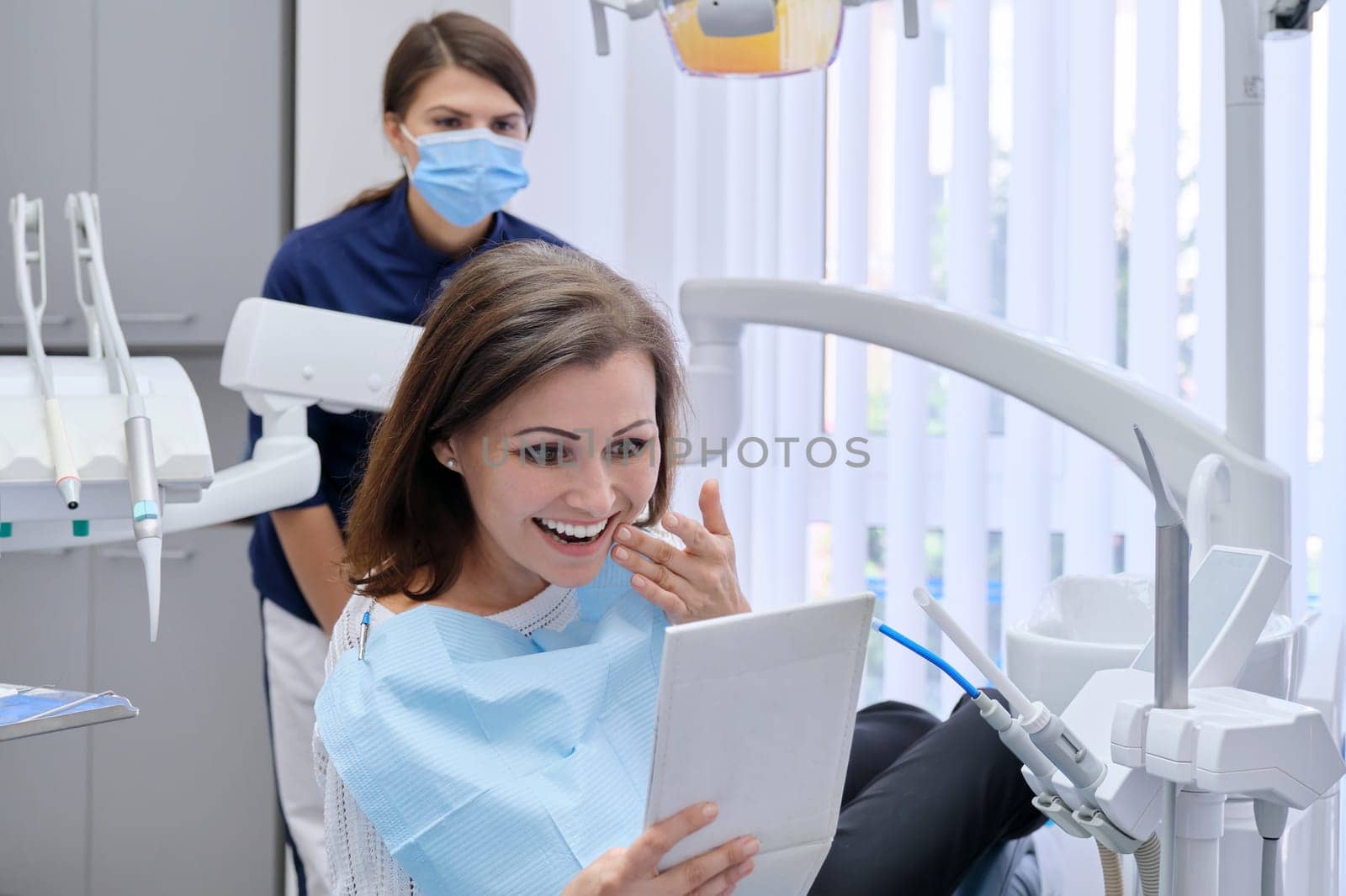 Happy middle aged woman with doctor dentist looking in mirror at teeth, sitting in dental chair. Medicine, dentistry and health care concept