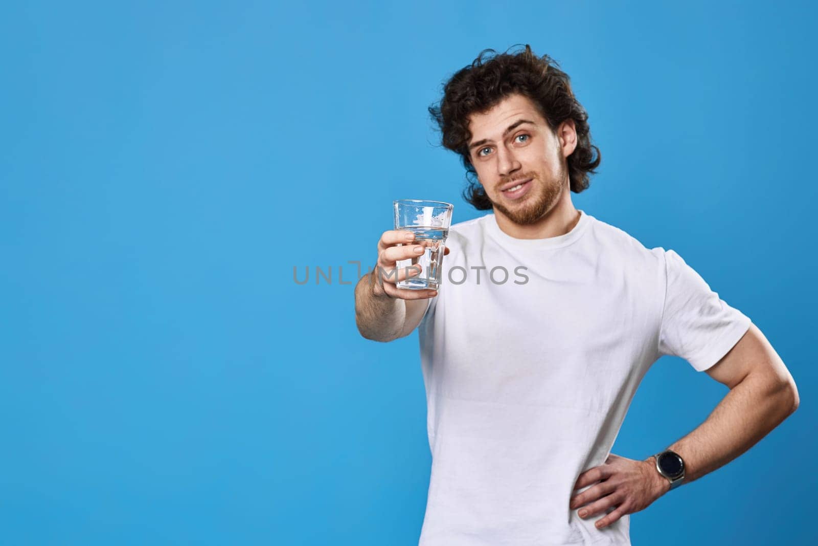 Young happy curly man showing drinking glass of water on blue background. focus on glass