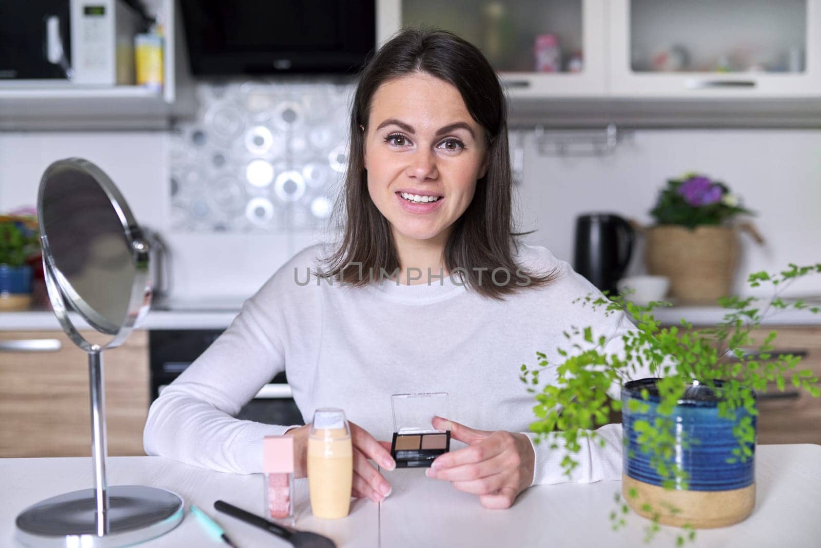 Young attractive woman doing makeup, female sitting at home at table in kitchen with cosmetic products mirror brush, shows eyebrow shadow makeup. Beauty, skin care, fashion, make up concept