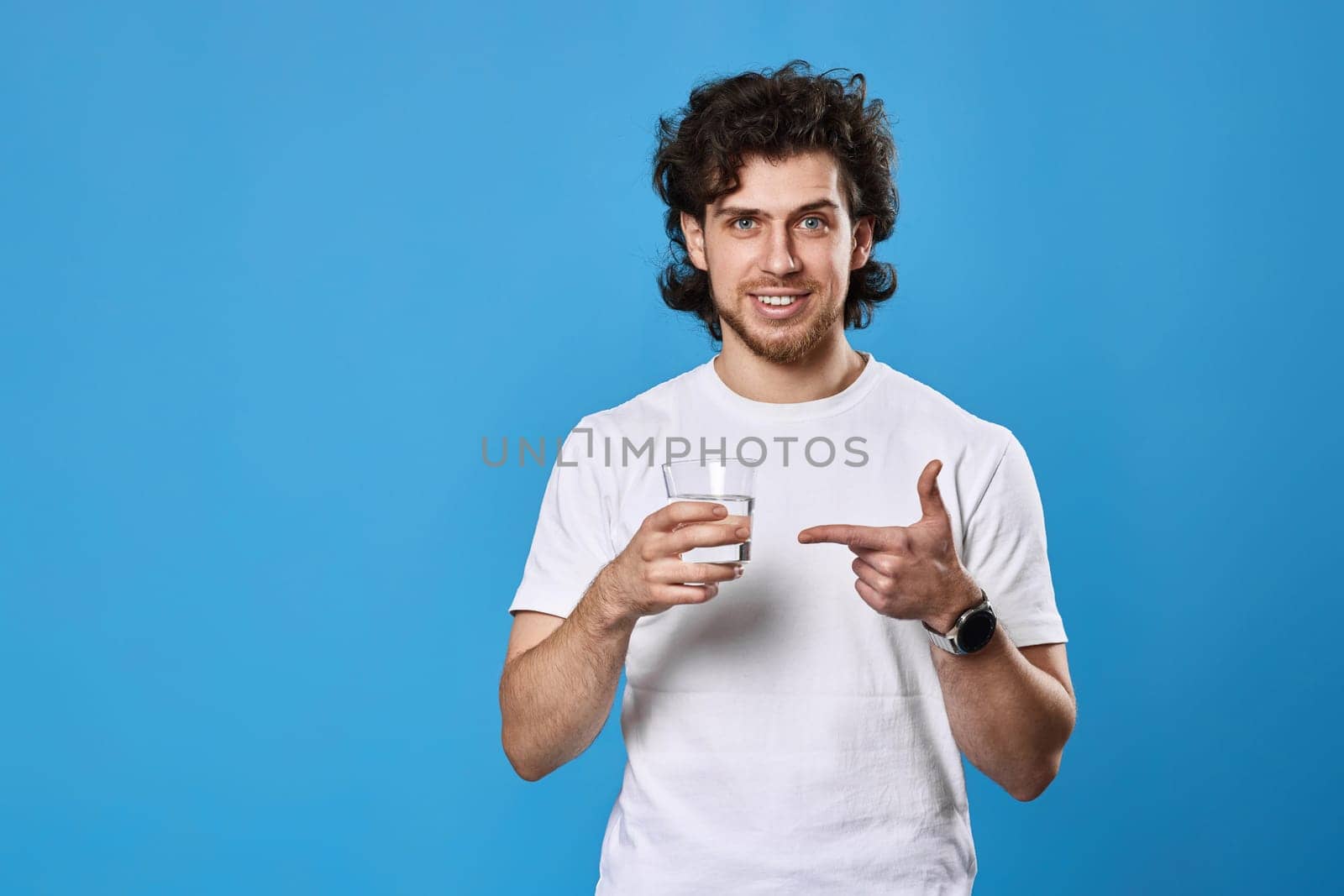 curly handsome man in white t-shirt showing drinking glass of water on blue background