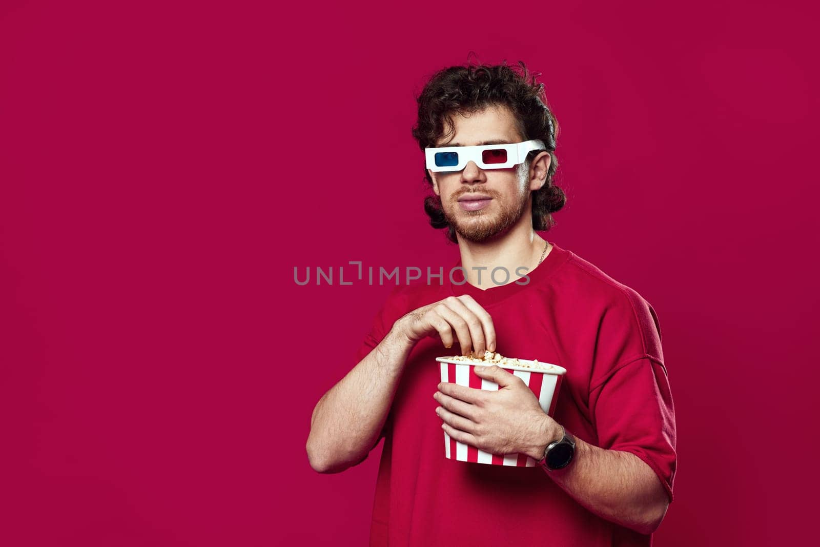 handsome bearded man in 3d glasses holding big bucket of popcorns on magenta background