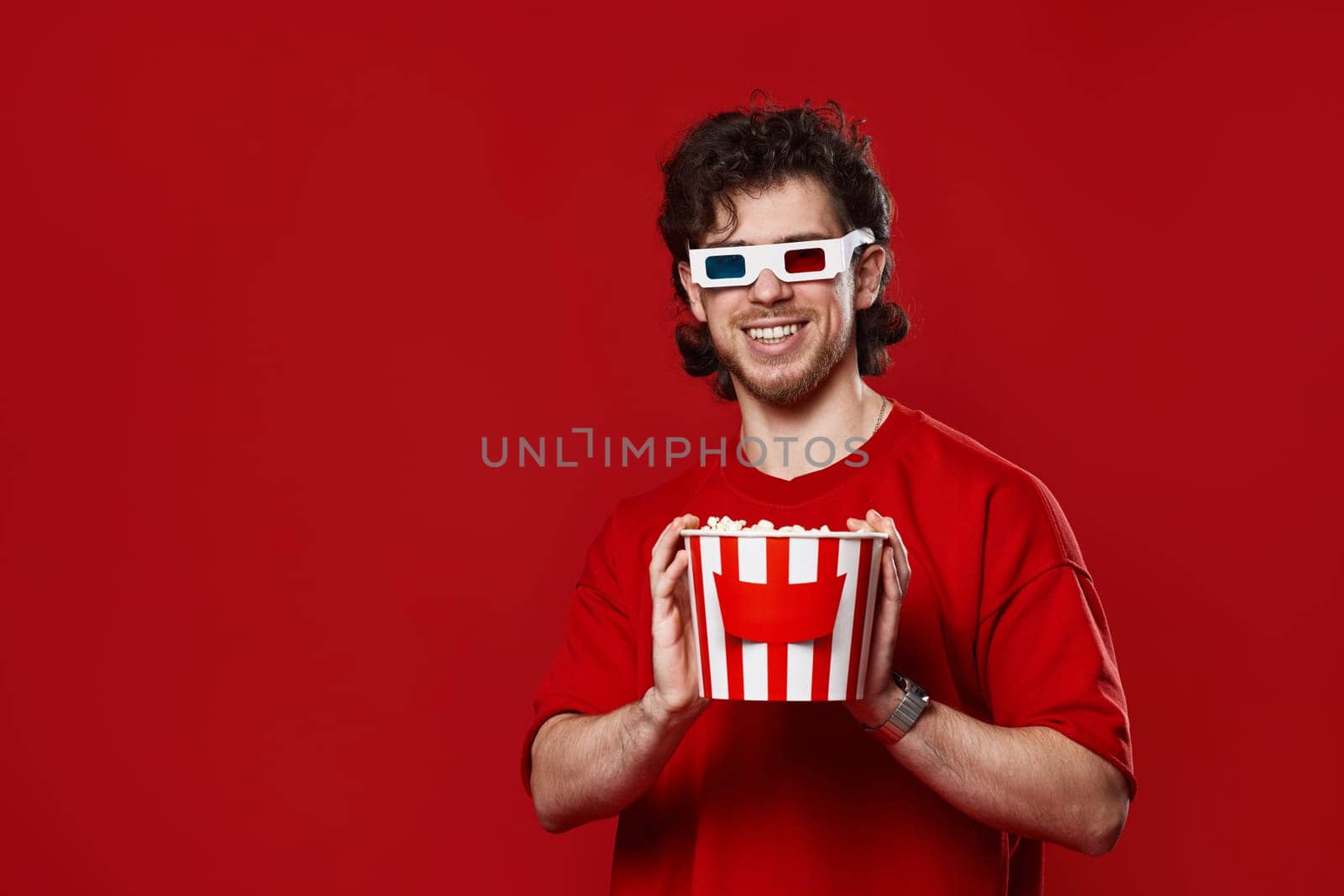 handsome bearded man in 3d glasses holding big bucket of popcorns on red background