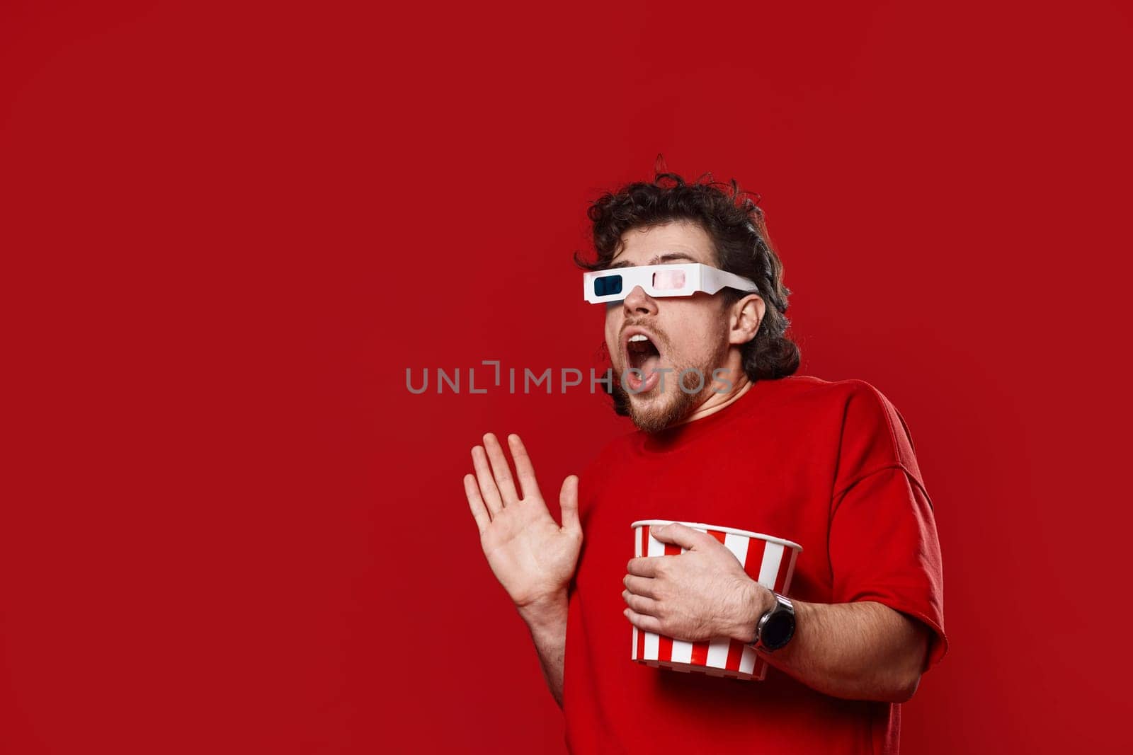 Shocked scared handsome man in 3d-glasses watching movie film hold bucket of popcorn on red background. copy space