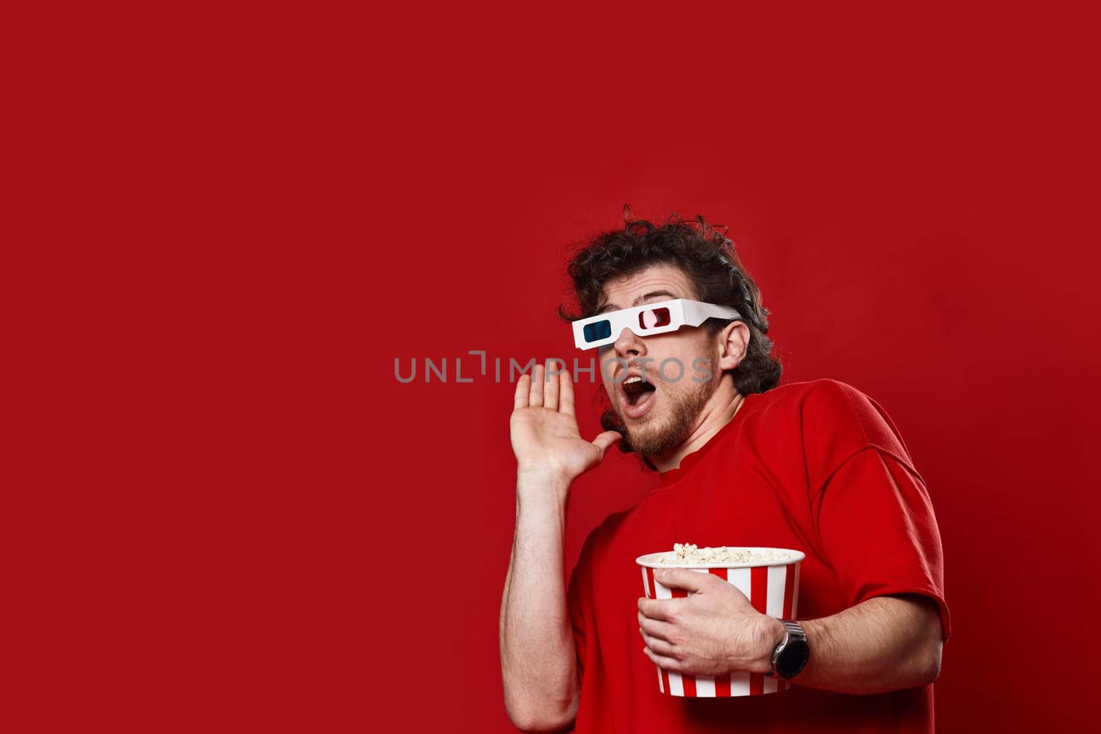 scared handsome bearded man in 3d-glasses holding bucket of popcorn on red background
