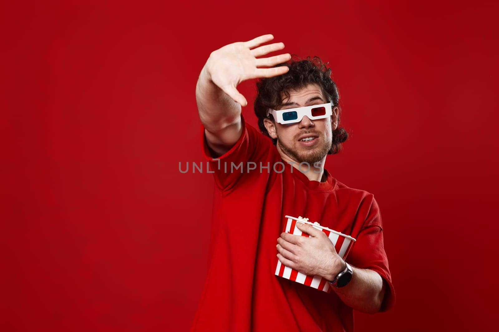 scared handsome man in 3d-glasses holding bucket of popcorn on red background