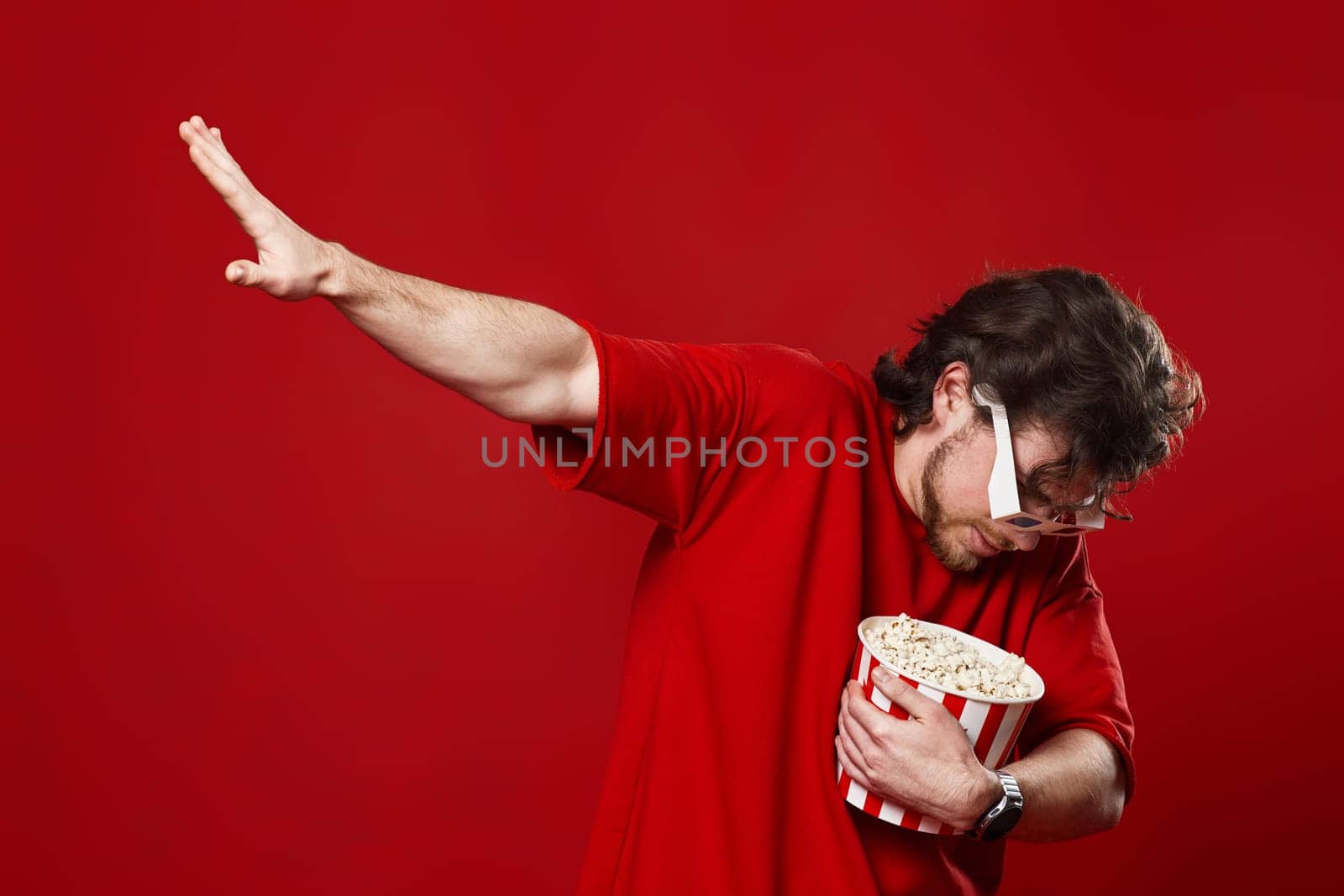 young bearded man in 3d-glasses holding big bucket of popcorn and dancing on red background