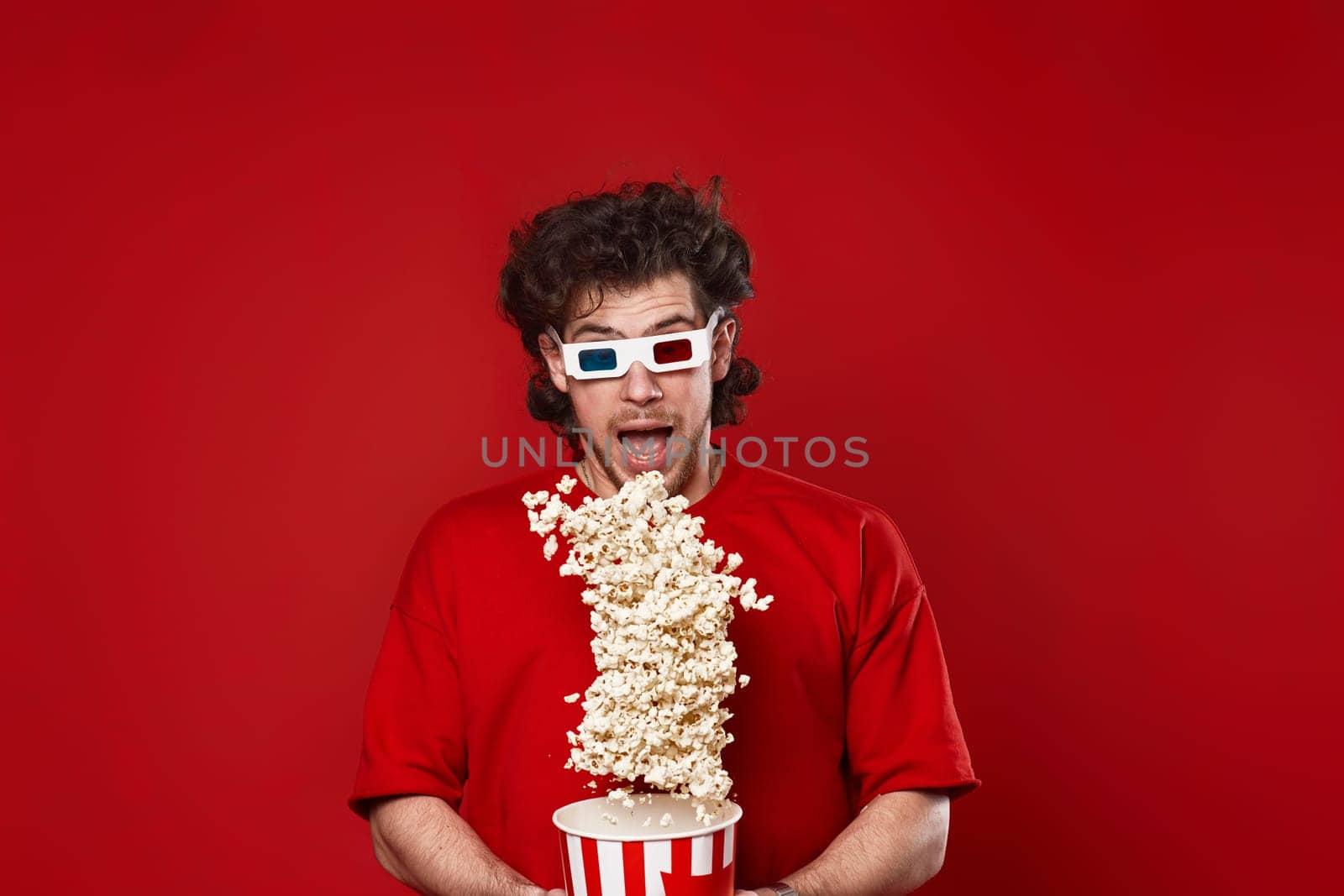 young handsome man in 3d glasses, red t-shirt jumping with big bucket of popcorn on red background