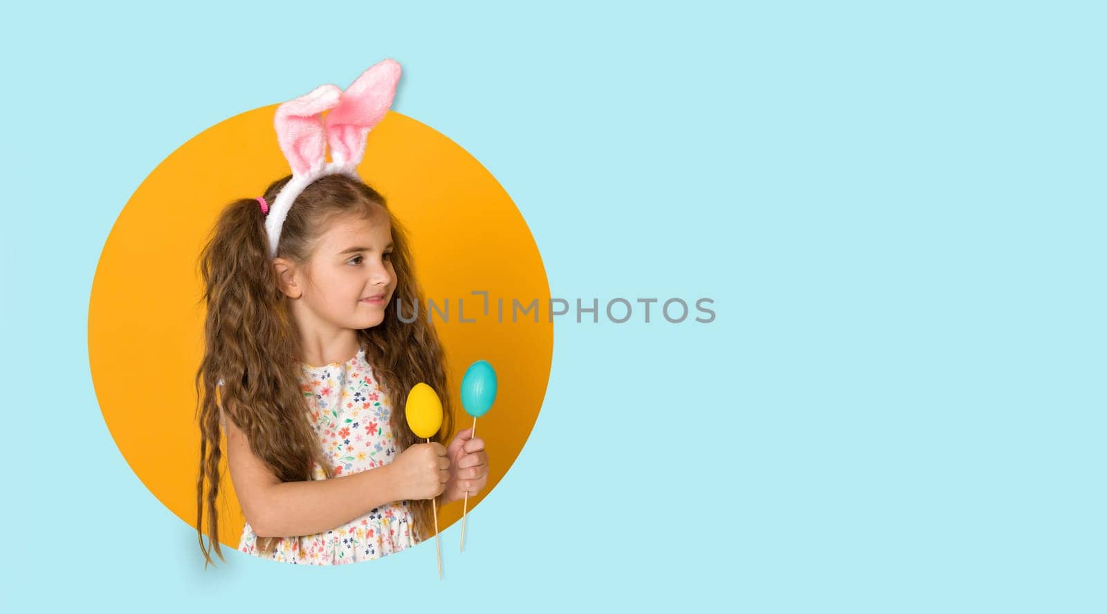 smiling curly caucasian little child girl with Easter bunny ears and colorful eggs on yellow and blue background. copy space