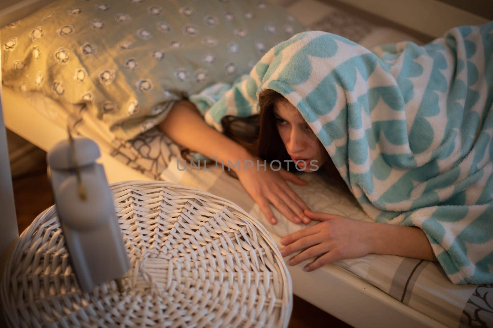 A frightened young woman hides under a blanket from the sound of her alarm clock. A girl under a soft blanket.