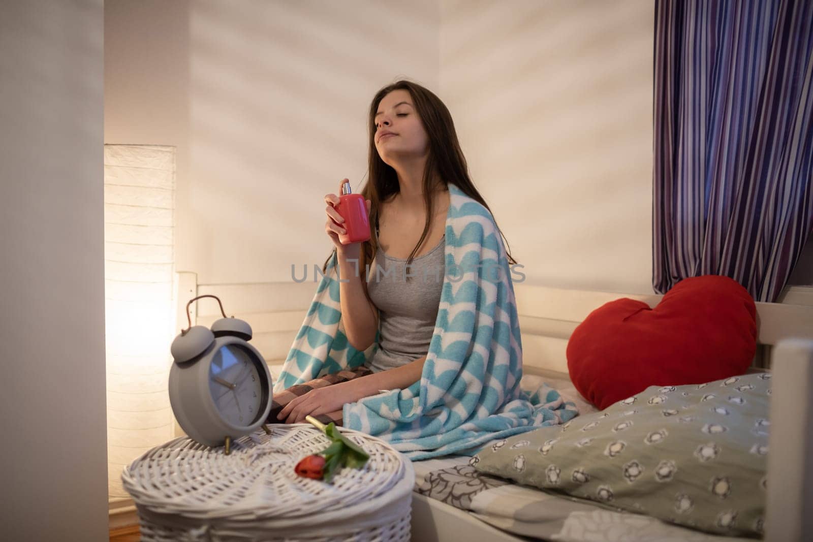 Girl holds pink perfume in her hands and sniffs it. A girl under a soft blanket.